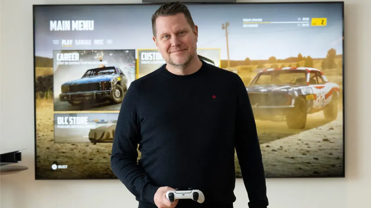 Carolin Wingefors ler mot kameran med en spelkontroll i handen, framför en TV som visar spelets huvudmeny med racerbilar.