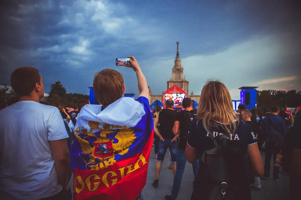 Młody kibic z flagą Rosji, wspierający klub piłkarski z Moskwy, filmuje telefonem wydarzenie przed monumentalnym budynkiem.