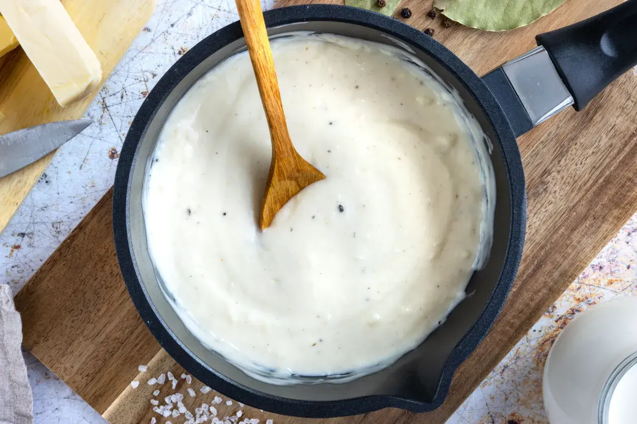 Cremige Bechamel-Sauce in einer Pfanne, bereit für die Lasagne. Butter, Milch und Gewürze sind daneben zu sehen.