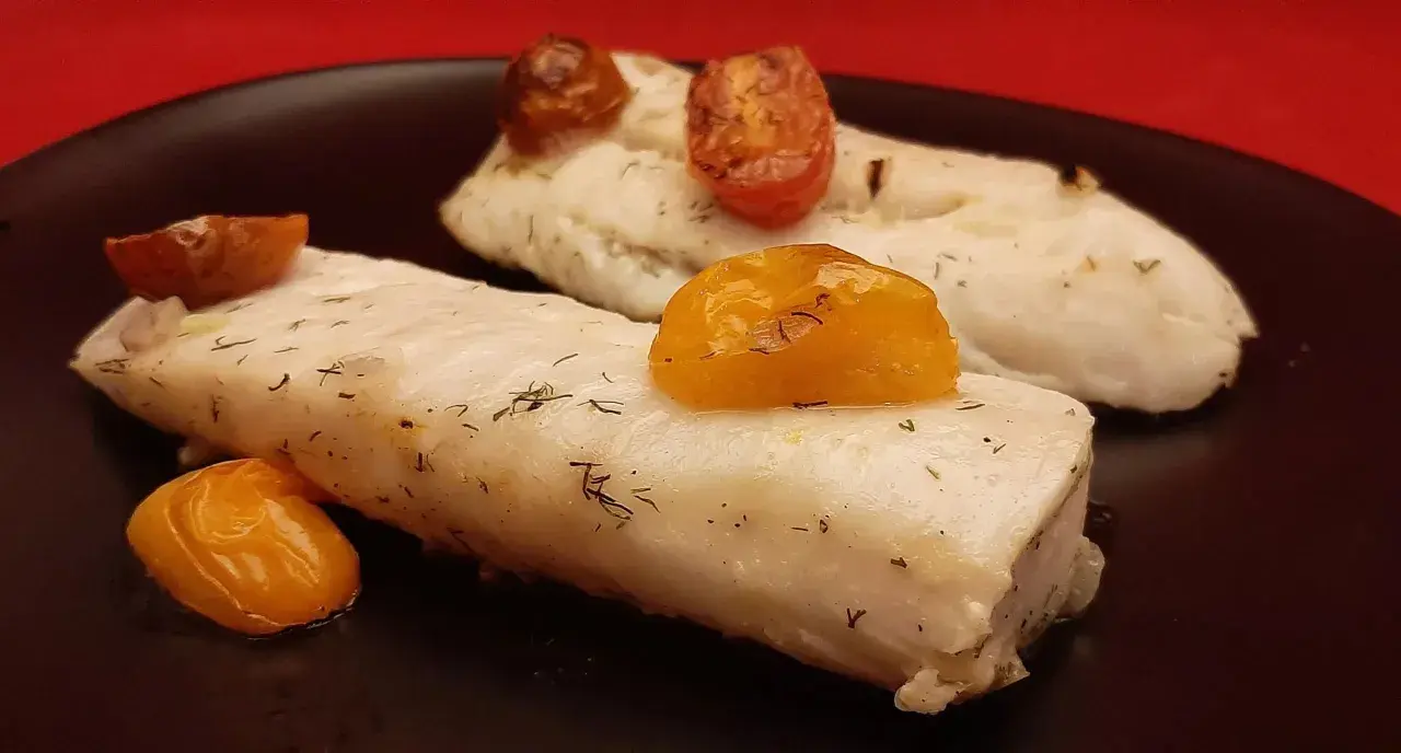 Deux pavés de dos de merlu tendres, parsemés d'aneth et surmontés de tomates cerises rôties, sur une assiette noire.