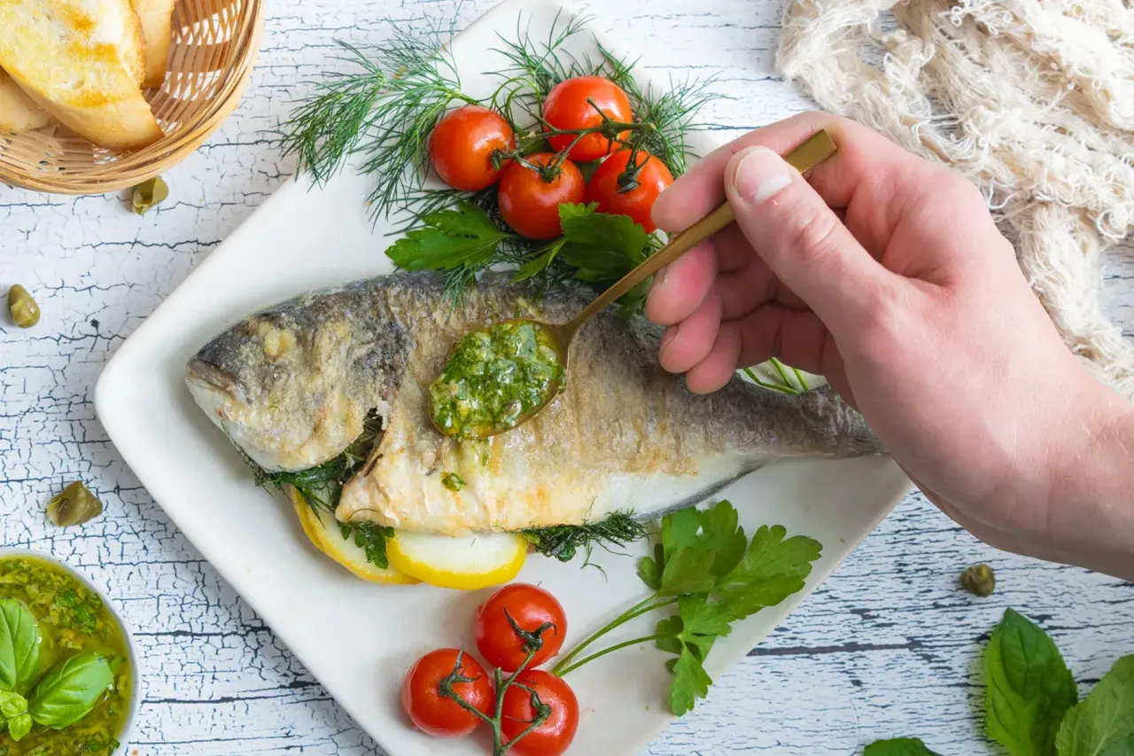 Eine goldbraune Dorade in der Pfanne, garniert mit frischen Kräutern, Kirschtomaten und Zitronenscheiben. Eine Löffelspitze grüner Sauce wird auf den Fisch gegeben.
