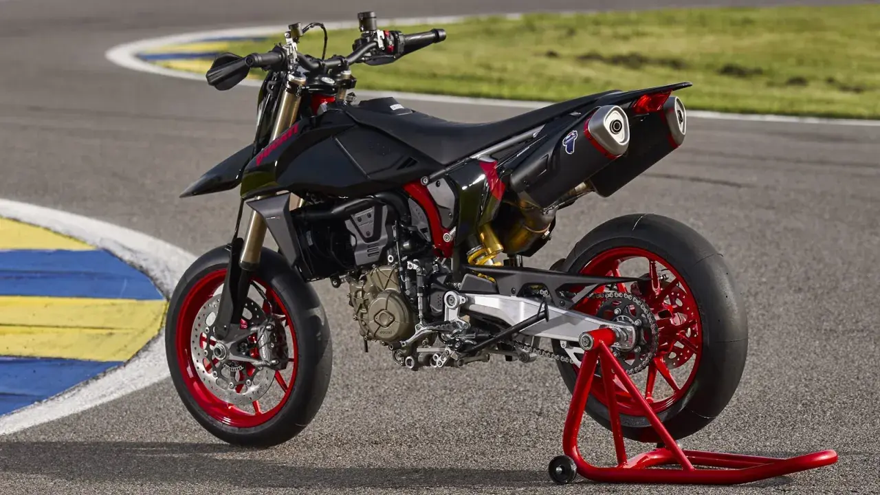 Czarny motocykl **hypermotard** z czerwonymi felgami i akcentami, stojący na czerwonym stojaku na torze wyścigowym.