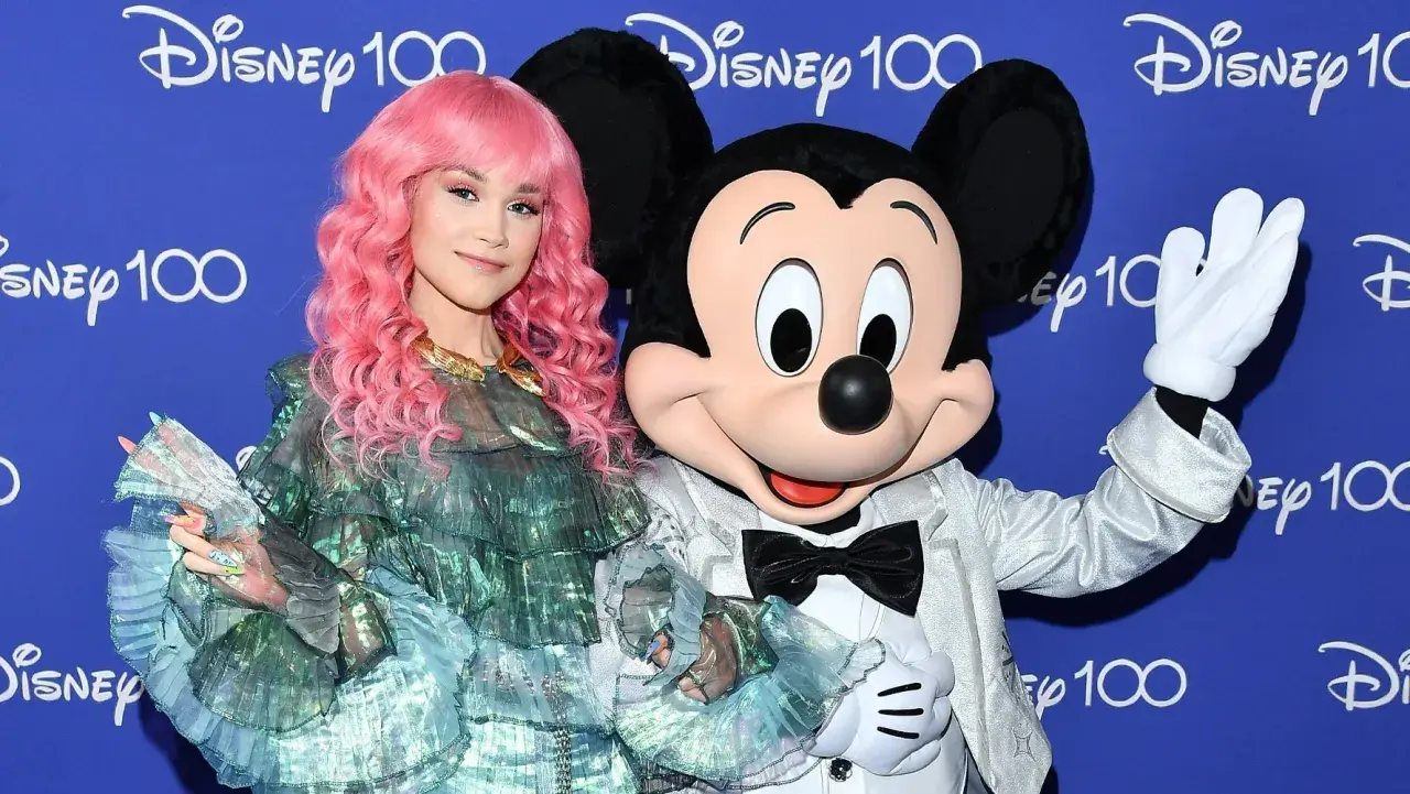 Dziewczyna z różowymi włosami i Myszka Miki pozdrawiają na tle napisu "Disney 100". Może to zapowiedź nowych piosenek Disneya?