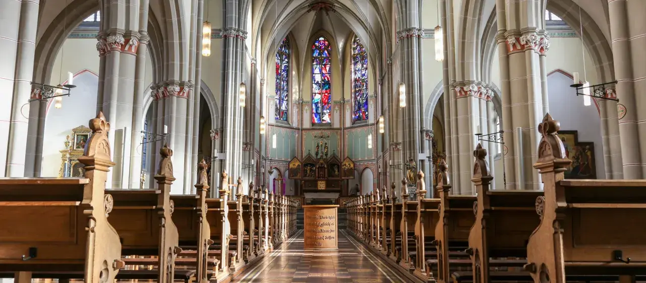 Blick in den katholischen Kircheninnenraum mit Holzbänken, Buntglasfenstern und einem Altar.