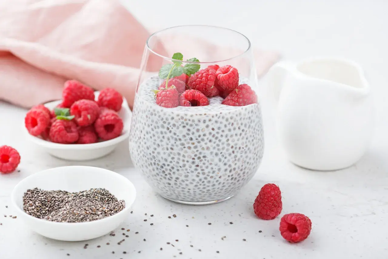 Pyszny deser chia pudding z malinami i miętą, obok miseczka z nasionami chia i świeżymi malinami.