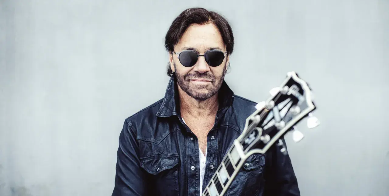 Al Di Meola w czarnej skórzanej kurtce i okularach przeciwsłonecznych, trzymający gitarę.