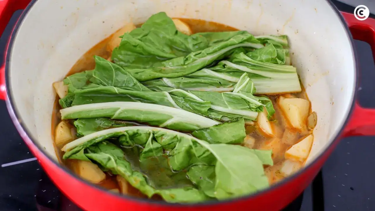 Verduras frescas, como acelgas, se cocinan en una olla roja con trozos de patata en un caldo sabroso.