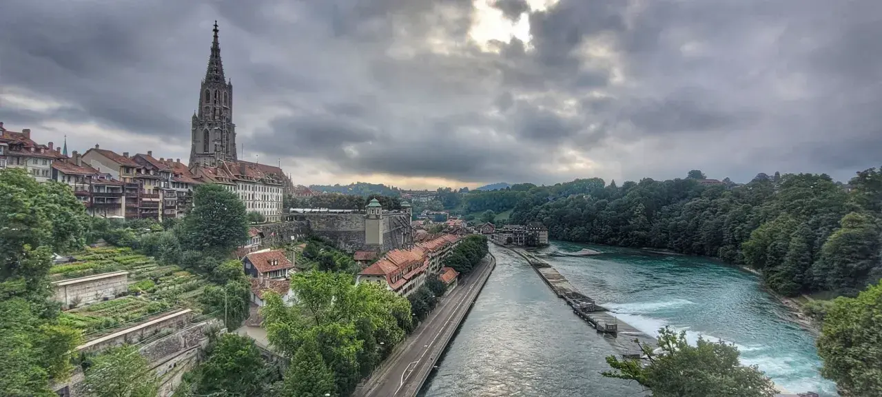 Blick von der Besucherplattform über Bern: Münster, Aare und grüne Hänge unter dramatischem Himmel.