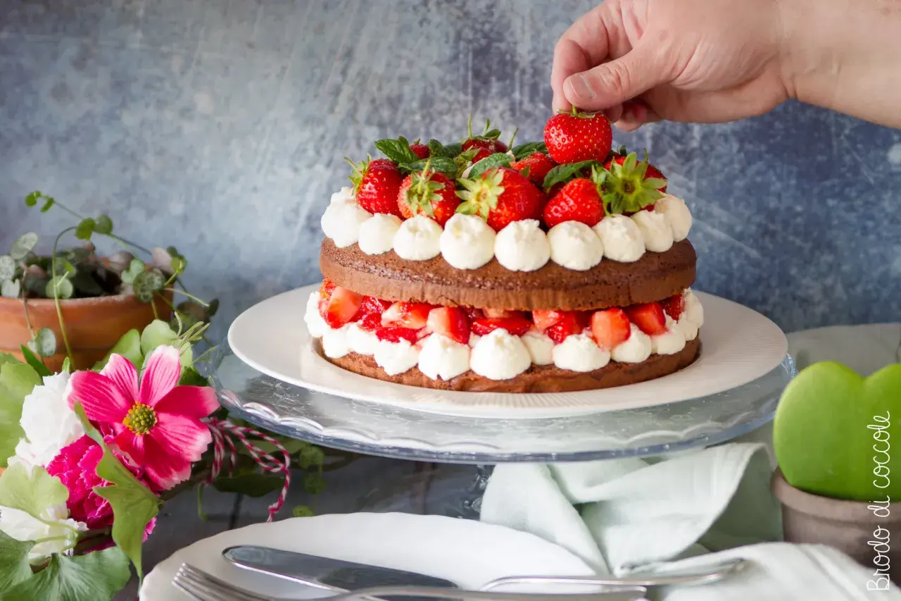 Una deliziosa torta con fragole e cioccolato, decorata con panna montata e fragole fresche. Una mano aggiunge l'ultima fragola in cima.