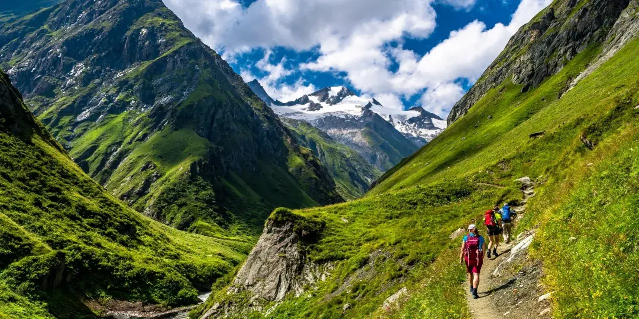 Hikers on a trail enjoying alpine hiking amidst lush green mountains and snow-capped peaks under a cloudy blue sky.