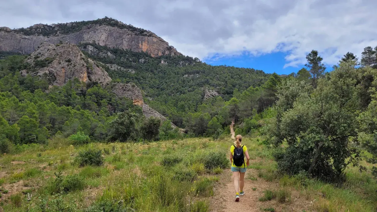 Senderista camina por un sendero en los **estrets d'Arnes**, rodeada de pinos y montañas rocosas bajo un cielo nublado.