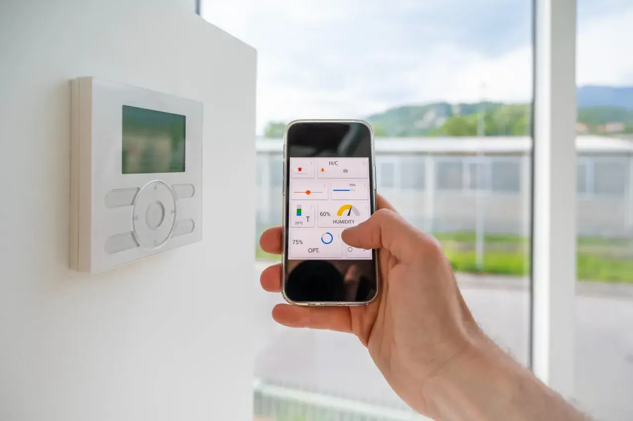 Control de la automatización de casas desde un smartphone. Se ve un termostato en la pared y una mano sujetando un teléfono con una app de control.