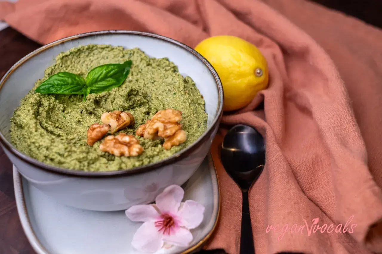 Un delicioso pesto vegano en un cuenco, adornado con hojas de albahaca y nueces. Junto a un limón y una flor rosa.