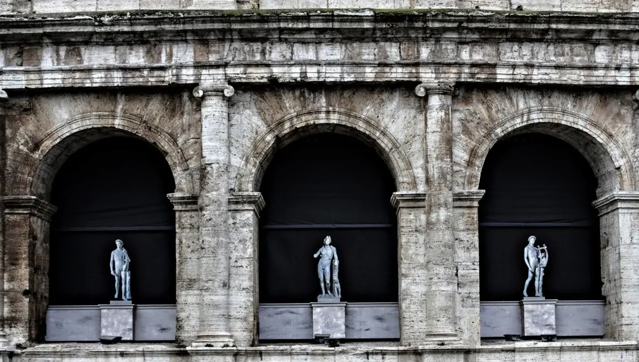 Tre statue classiche emergono dall'ombra di un imponente arco a Roma, testimoni silenziose di un passato glorioso.