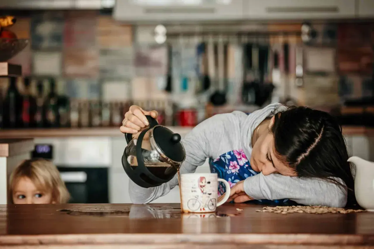 Une femme, visiblement à bout de forces, verse du café dans une tasse, le regard perdu. Un enfant la regarde, l'air intrigué. Elle semble tomber de fatigue.
