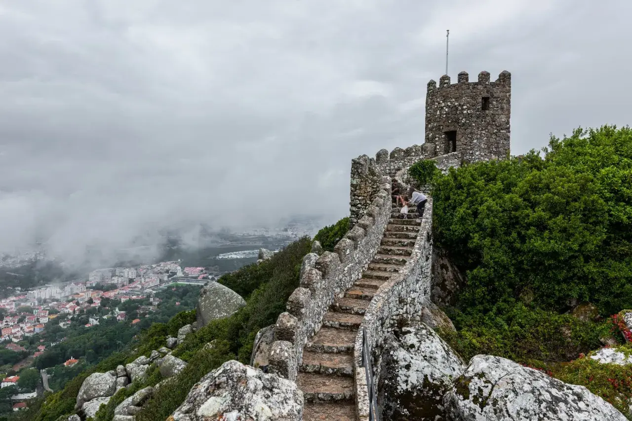 Escaleras de piedra ascienden hacia una torre en el **castillo de Sintra**, con vistas a la ciudad envuelta en niebla.