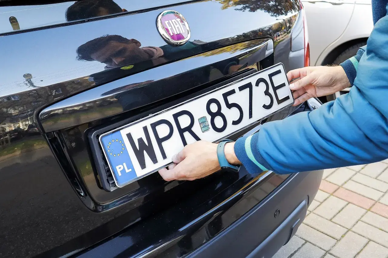 Ręce montują tablicę rejestracyjną WPR 8573E na czarnym samochodzie. To duplikat tablicy rejestracyjnej.