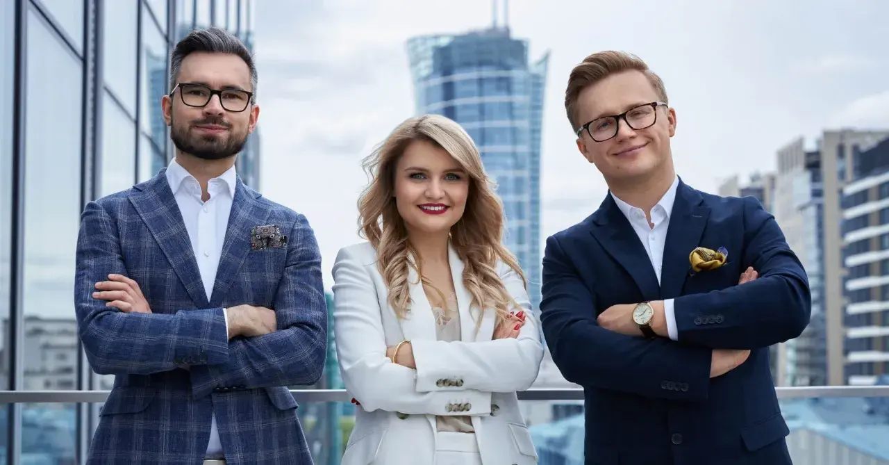 Trzy osoby w eleganckich garniturach stoją na tle nowoczesnych budynków.