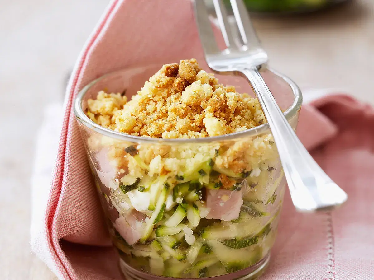 Un délicieux crumble de courgettes, jambon et fromage, servi dans un verre, prêt à être dégusté avec une fourchette.