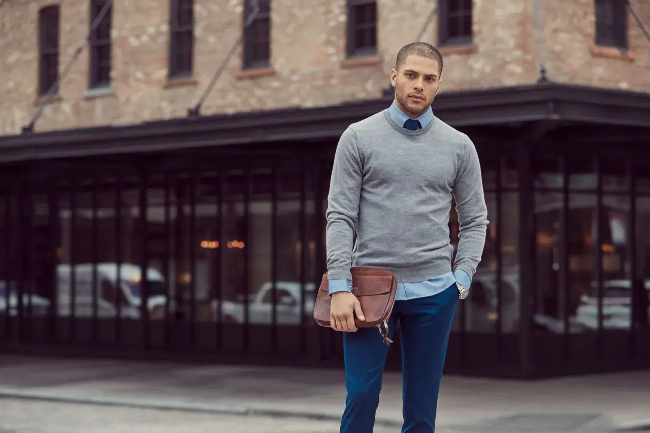 Ein Mann mit Stil, der sein **Outfit zusammenstellen Herren** perfektioniert: grauer Pullover, hellblaues Hemd, blaue Hose und eine braune Ledertasche.