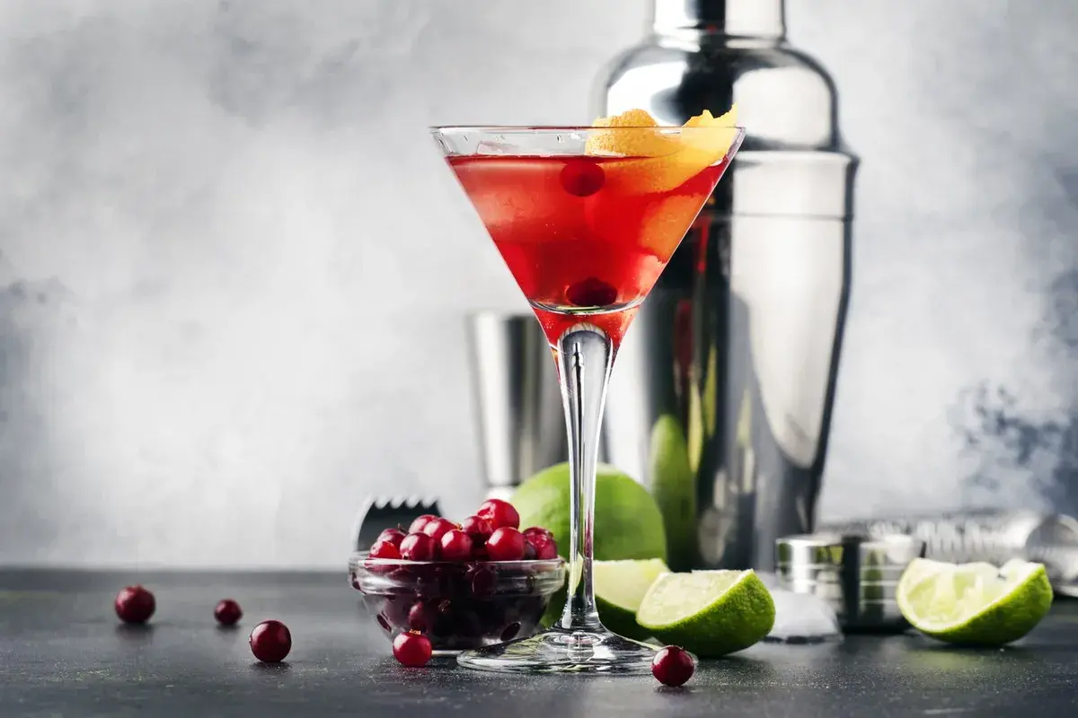 Elegancki Cosmopolitan przepis: czerwony drink w kieliszku, obok żurawina, limonka i shaker. Idealny na wieczór.