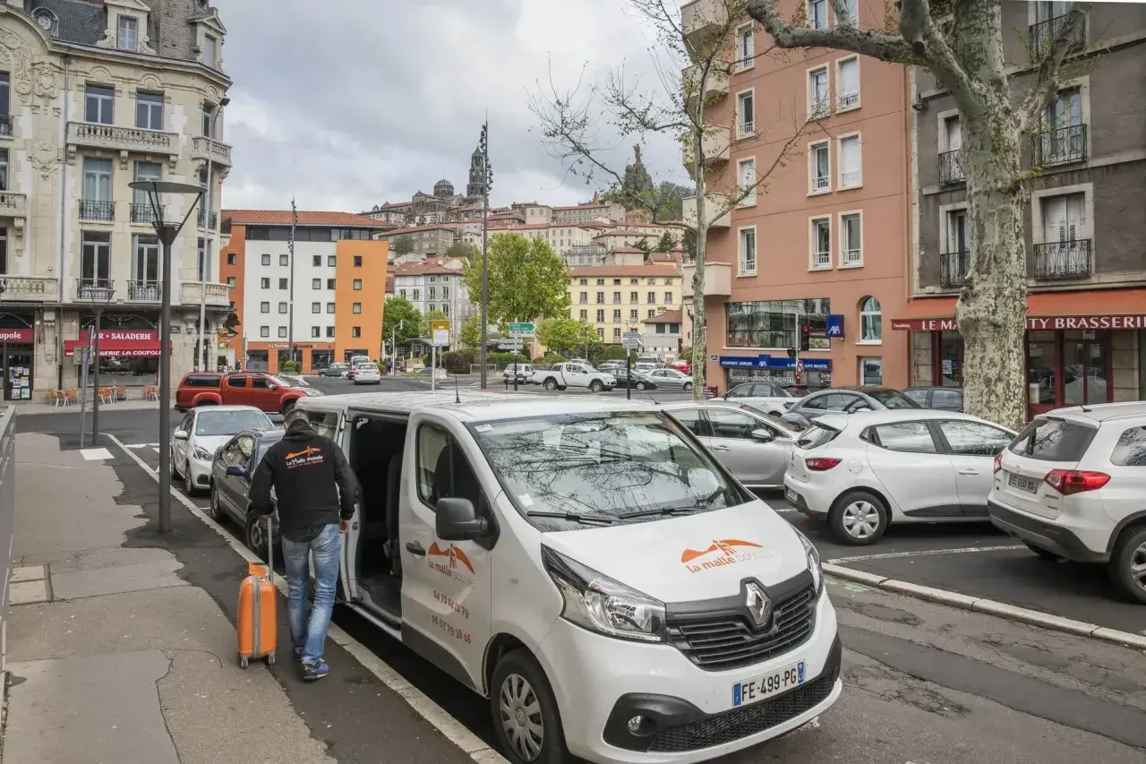 Un homme charge une valise orange dans un bus compostelle blanc. La ville s'étend en arrière-plan.