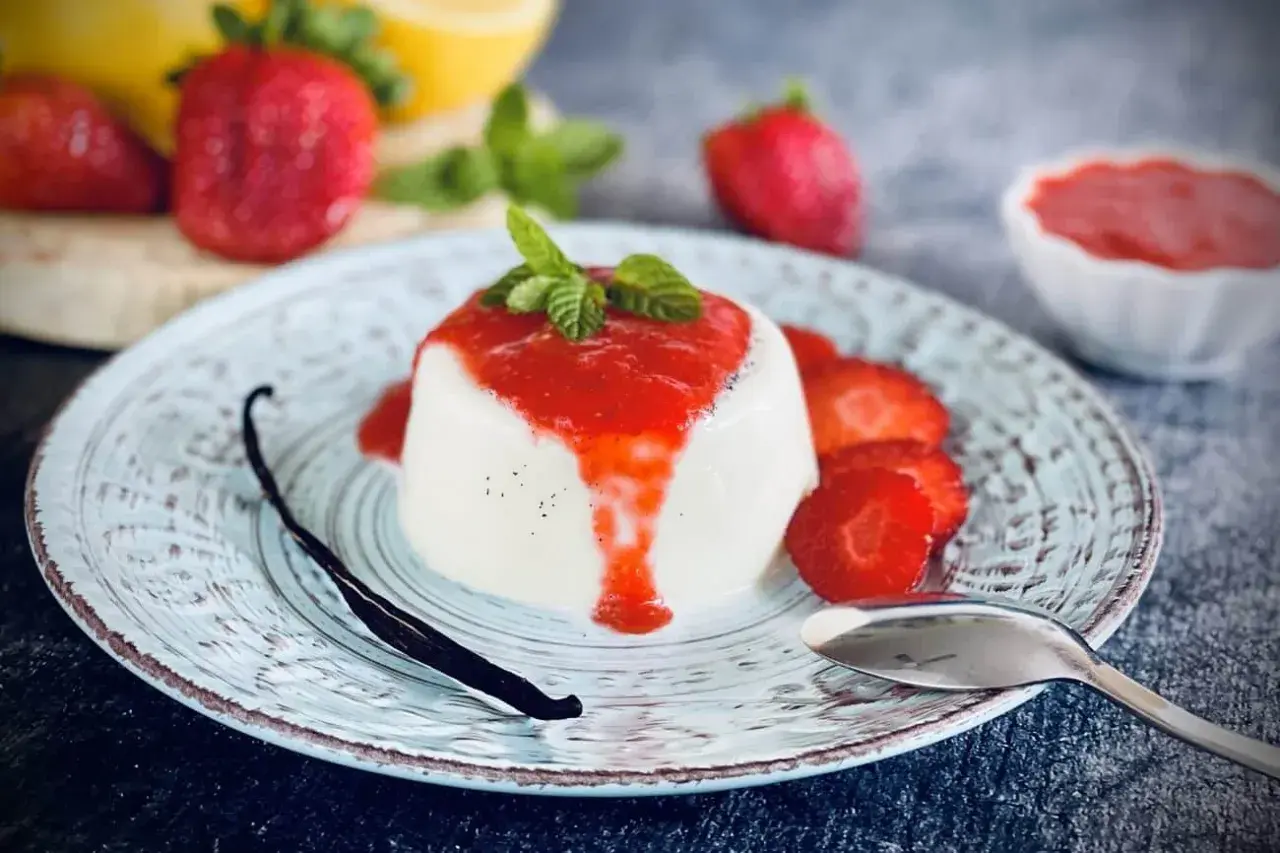 Cremige Panna Cotta mit Erdbeersauce, frischen Erdbeeren, Minze und einer Vanilleschote auf einem Teller.