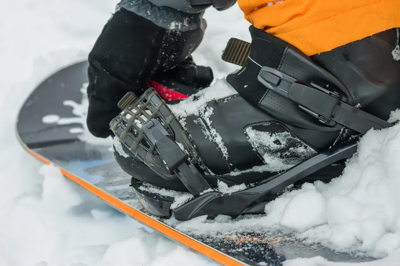 Rękawica zapina but snowboardowy na desce, przygotowując się do wygrzewania butów snowboardowych w śniegu.