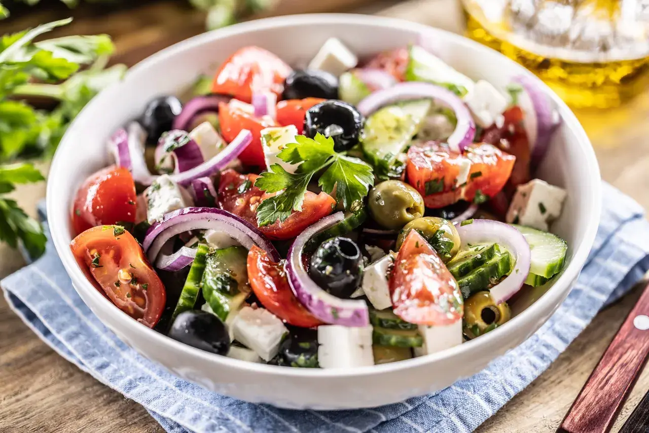 Ensalada griega fresca con tomates, pepinos, cebolla morada, aceitunas negras y queso feta, servida en un bol blanco.