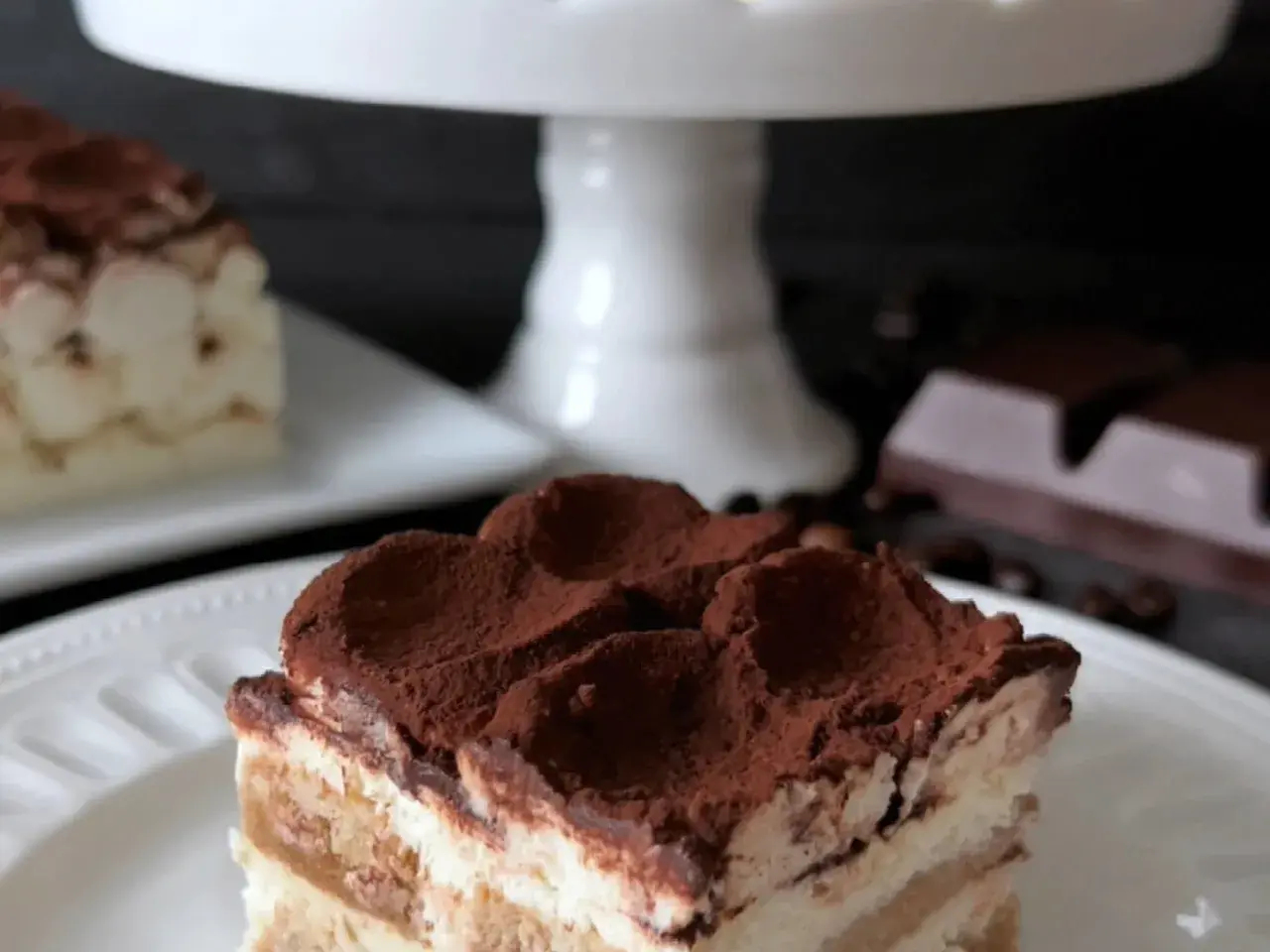 En bit krämig tiramisu, perfekt för ett tiramisu recept original. Pudrad med kakao, med kaffebönor och choklad i bakgrunden.