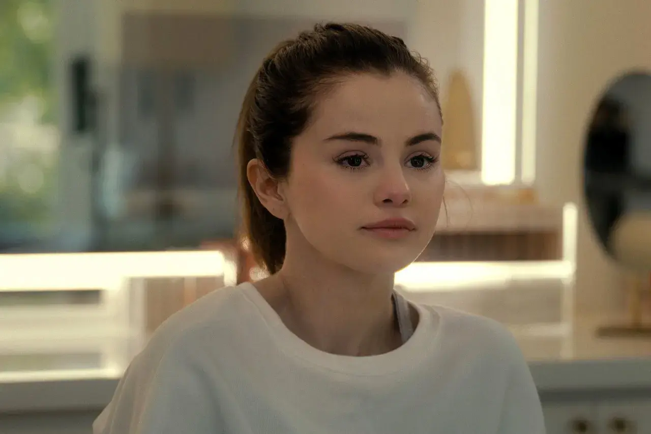 Selena Gomez w białej bluzie, z włosami związanymi w kucyk, patrzy w bok zamyślona.