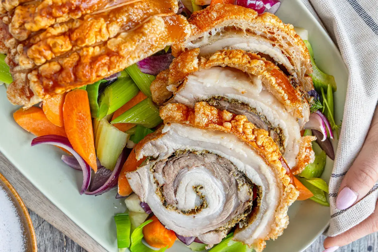 Knusprige Porchetta, ein italienischer Schweinebraten, wird mit karamellisierten Karotten, Sellerie und roten Zwiebeln serviert.