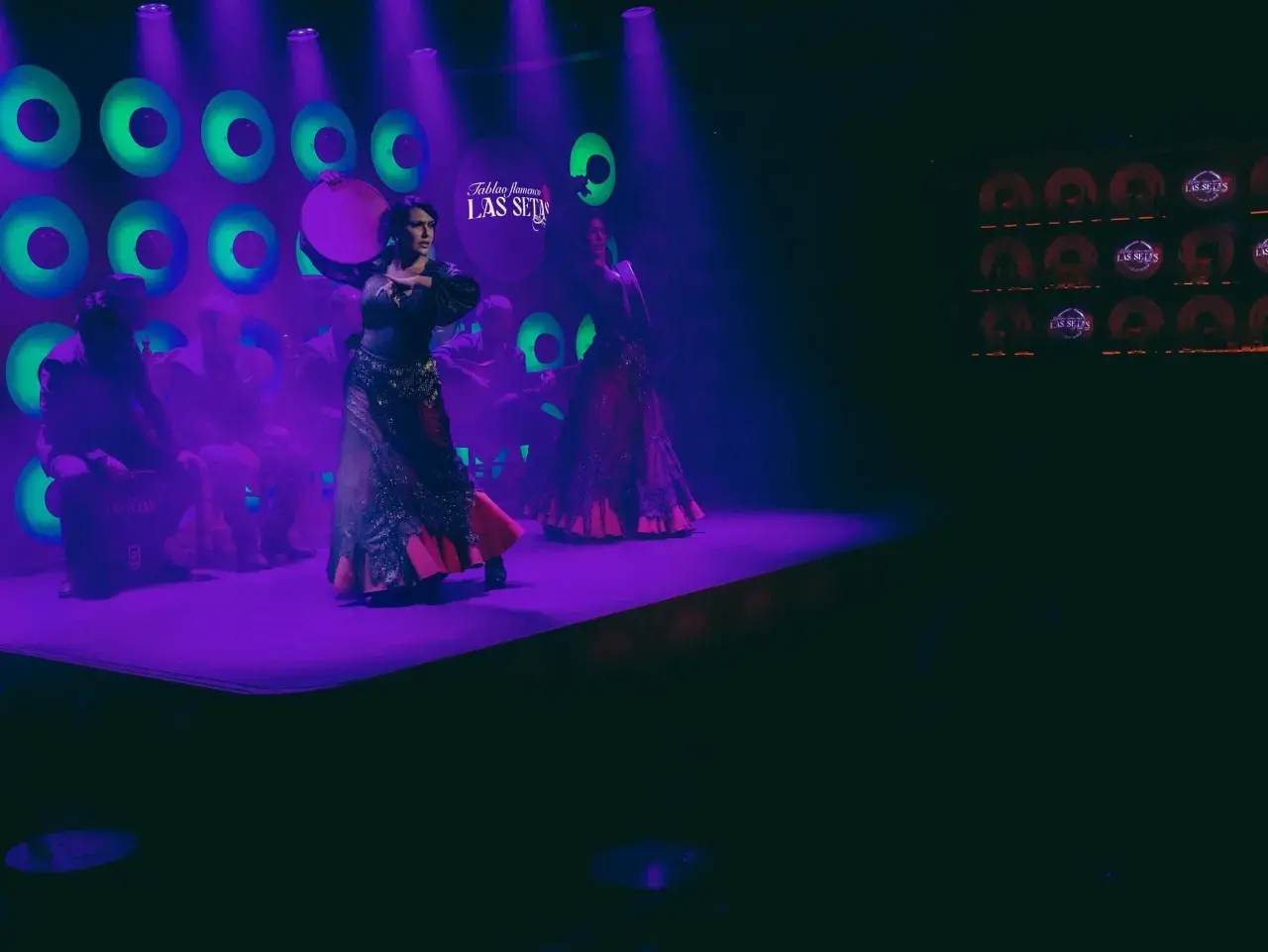 Dos bailarinas de flamenco, una con un tambor, aprenden a bailar sevillanas en un escenario con luces moradas y verdes.