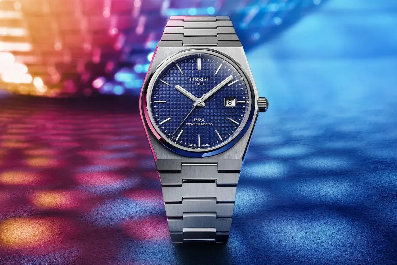 Reloj Tissot 1853 PRX Powermatic 80 con esfera azul texturizada y brazalete metálico, sobre un fondo vibrante de luces. Descubre su tissot 1853 precio.