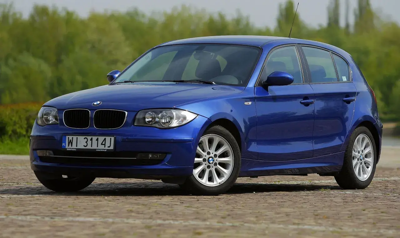 Niebieskie BMW E87 z polskimi tablicami WI 3114J, z nowymi żarówkami w reflektorach, zaparkowane na bruku.