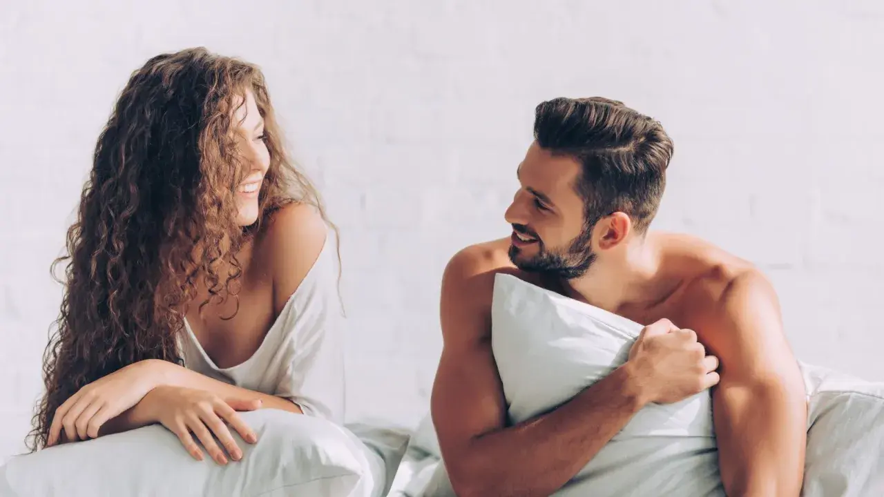 Coppia sorridente a letto, con la donna dai capelli ricci che guarda l'uomo con barba, un momento intimo prima di fare sesso.