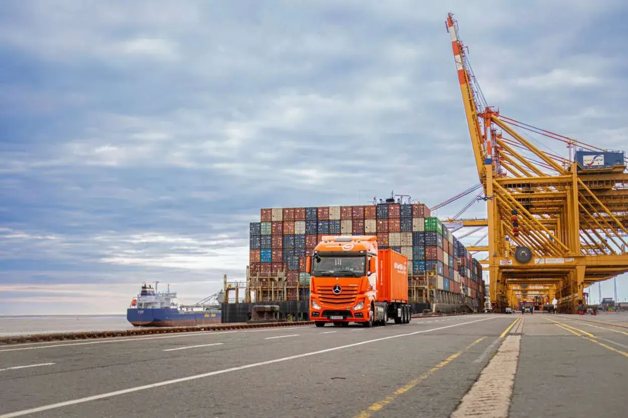 Ein orangefarbener LKW fährt an einem Containerschiff und einem Hafenkranausleger vorbei. Das zeigt, was eine Spedition leistet: Güter transportieren.
