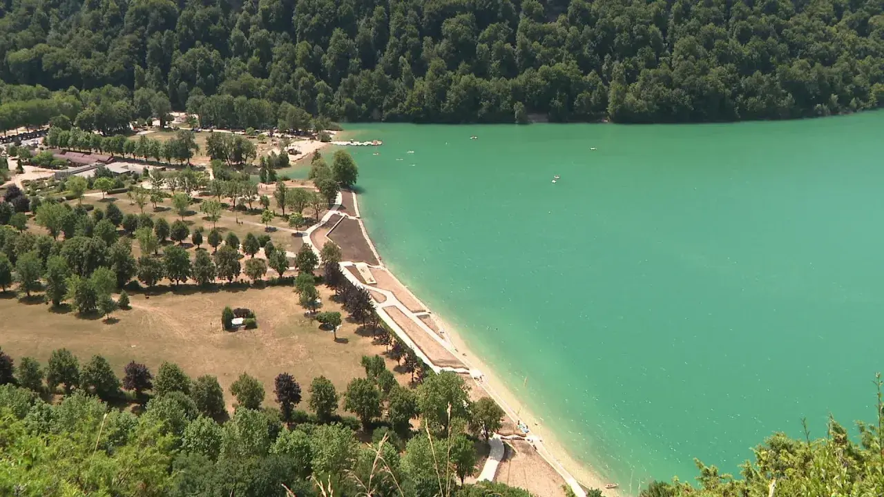 Vue aérienne d'un magnifique lac aux eaux turquoise, idéal pour la baignade. Des plages aménagées bordent la rive, entourées de verdure et d'arbres.