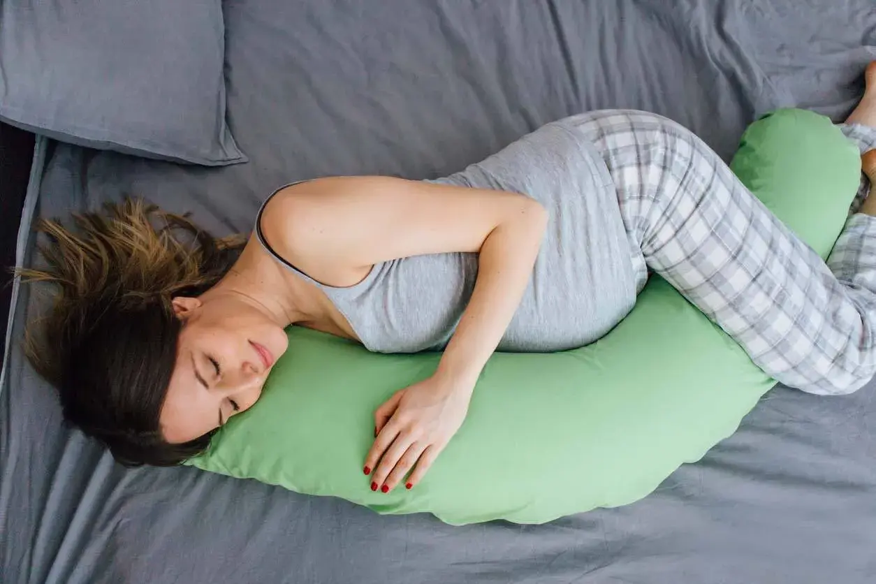 Donna incinta dorme su un fianco, supportata da un cuscino verde. Scopri come dormire in gravidanza per un riposo sereno.