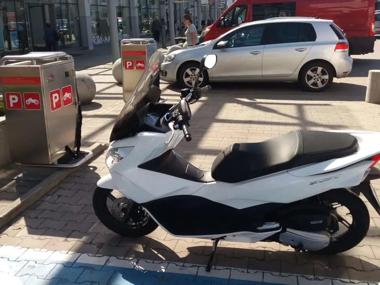 Na parkingu stoi biały skuter. Obok znajduje się automat z oznaczeniami "P" i symbolem motocykla, co sugeruje, że motocykle płacą za parking.