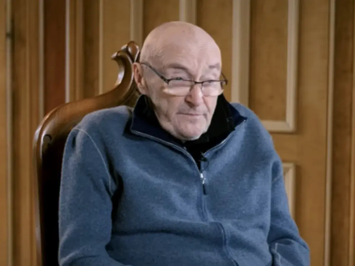 Phil Collins w okularach, siedzi w fotelu. Jego choroba wpływa na jego zdolność do gry na perkusji, ale nadal inspiruje fanów.