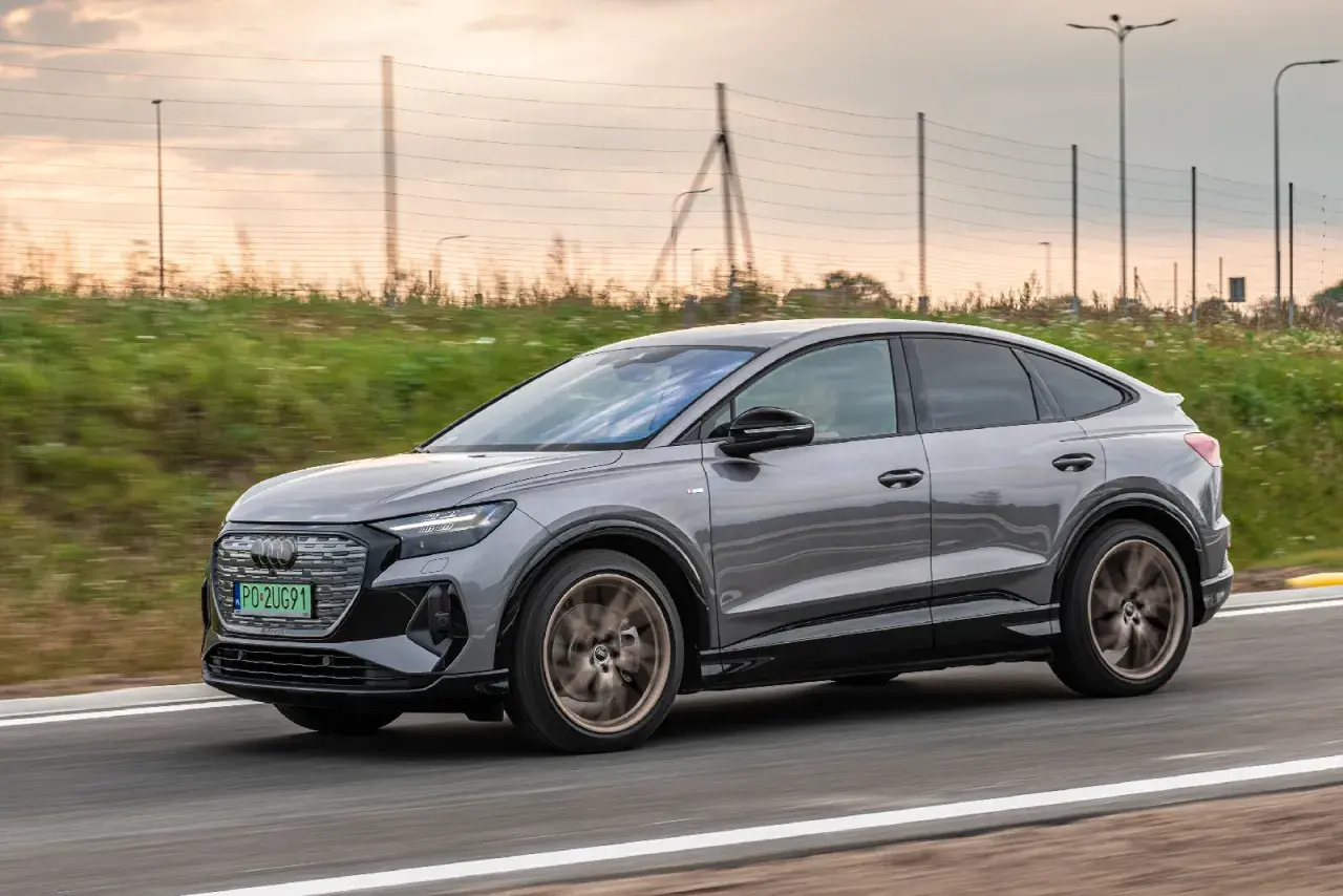 Szary Audi Q4 Sportback sunie po drodze, otoczony zielenią i zachodzącym słońcem.