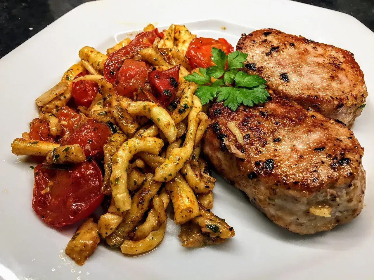 Deux côtelettes de veau grillées, servies avec des pâtes torsadées et des tomates cerises poêlées, garnies de persil frais. Un plat savoureux et coloré.