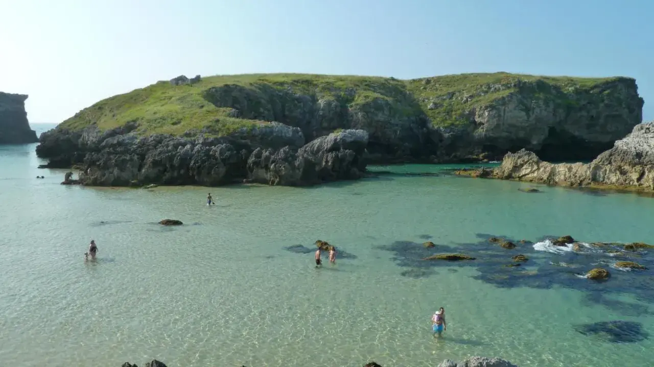 Gente disfrutando del agua cristalina en la costa de Llanes, con acantilados verdes y rocosos de fondo.