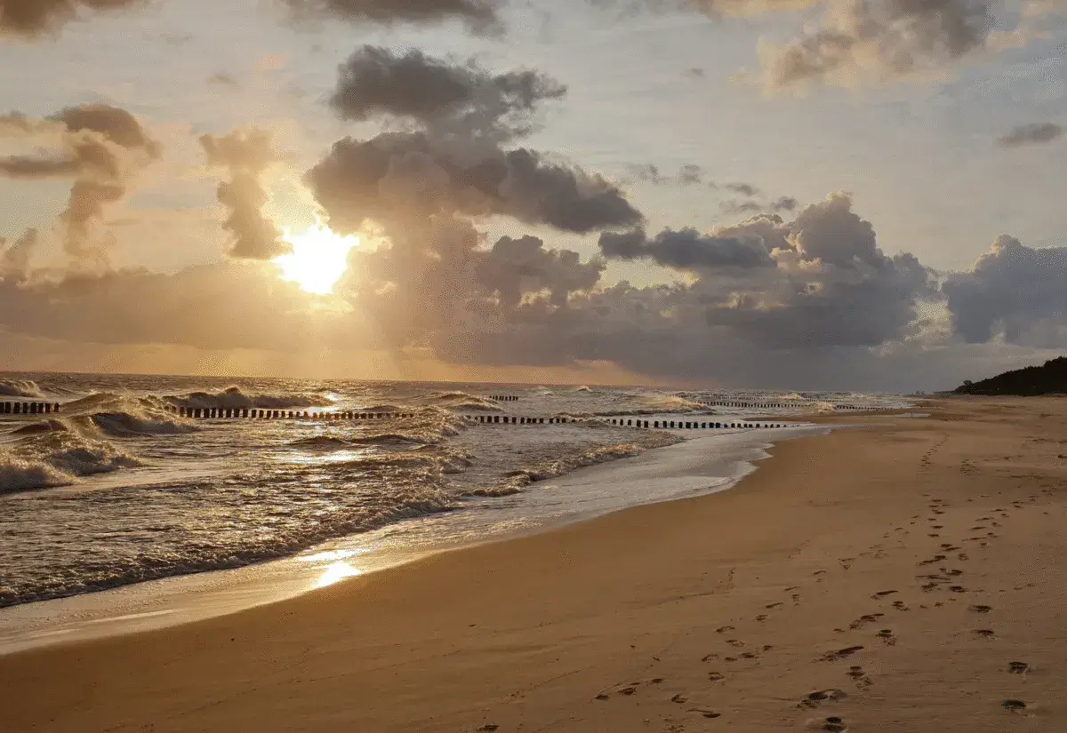 Piękny zachód słońca we Władysławowie. Słońce przebija się przez chmury, tworząc złote refleksy na falach Bałtyku. Na piaszczystej plaży widać ślady stóp.