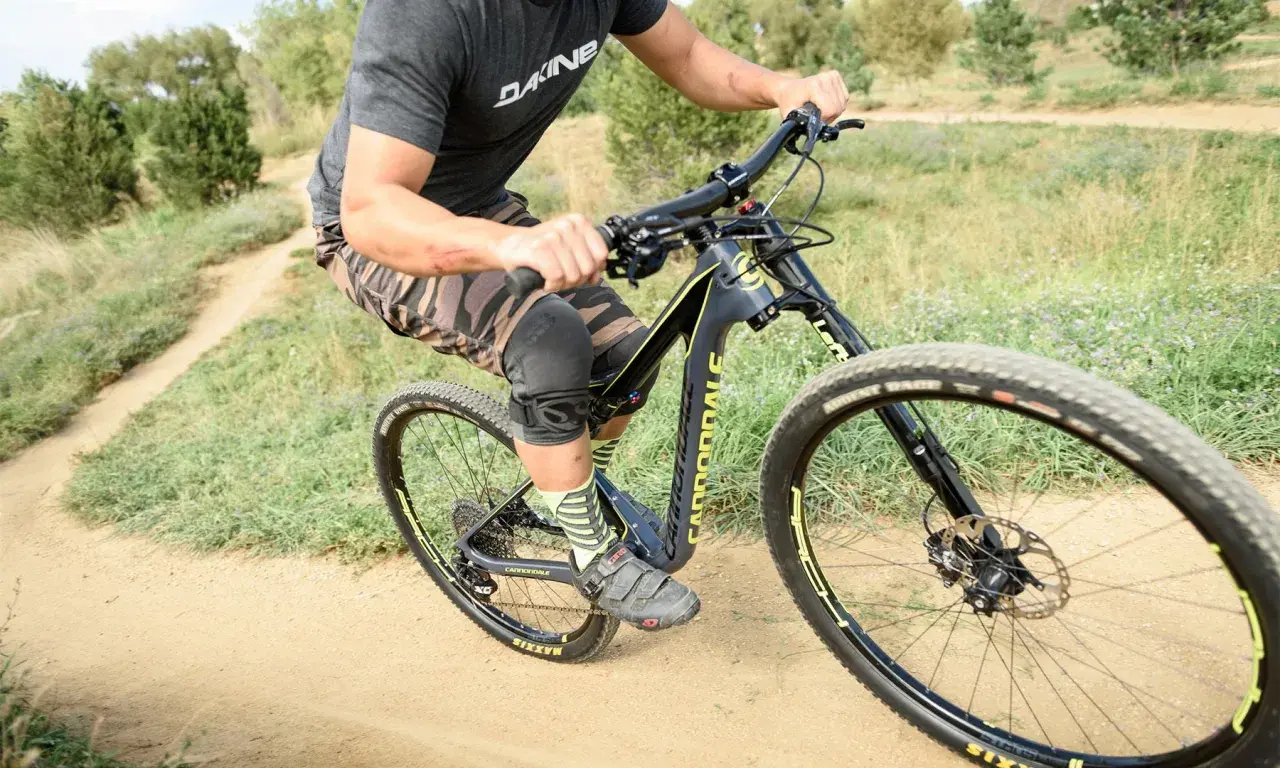 A rider on a black and yellow single fork mountain bike navigates a dusty trail, wearing knee pads and camouflage shorts.
