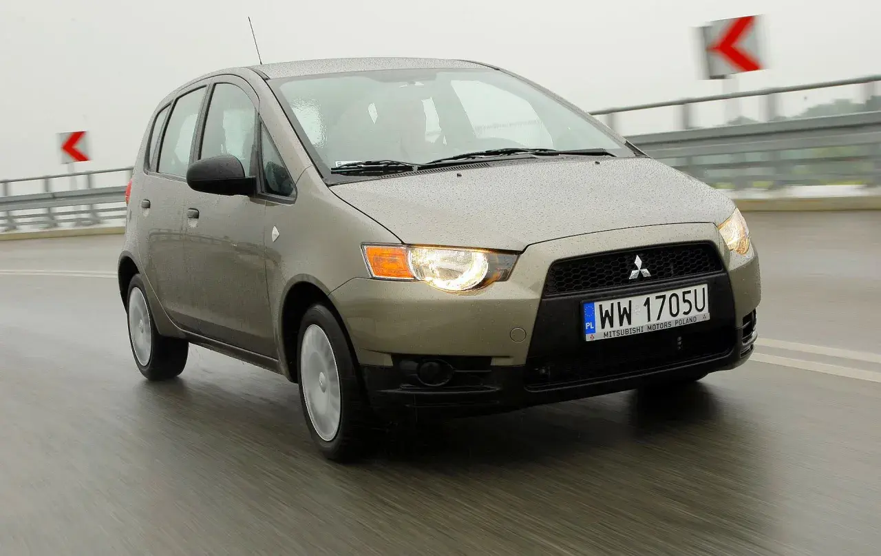 Złoty Mitsubishi Colt 2006 na mokrej drodze, światła włączone, w tle znaki drogowe.