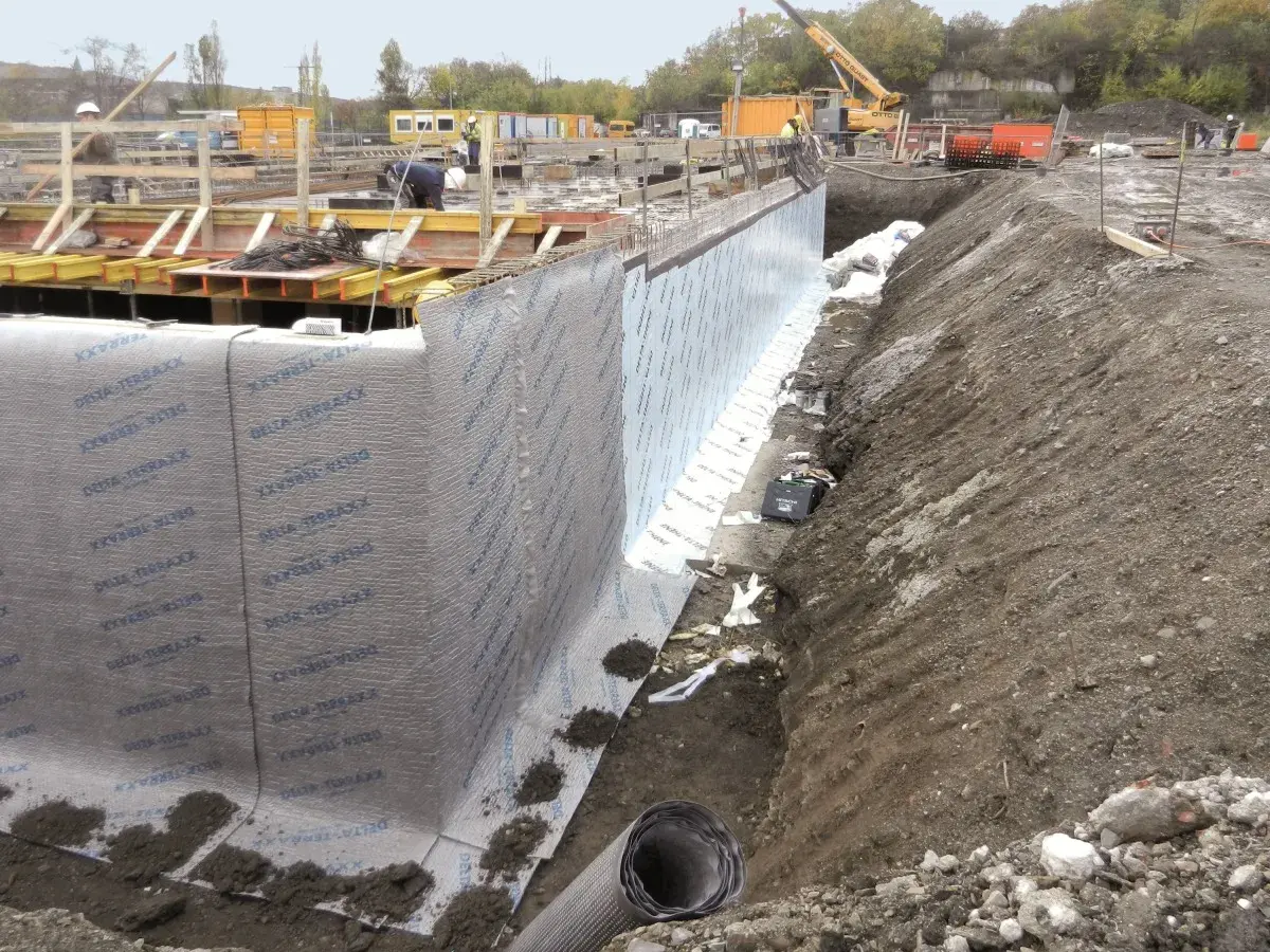 Baustelle mit frisch verlegter Noppenbahn wie rum. Betonfundament und Baugrube sind sichtbar.