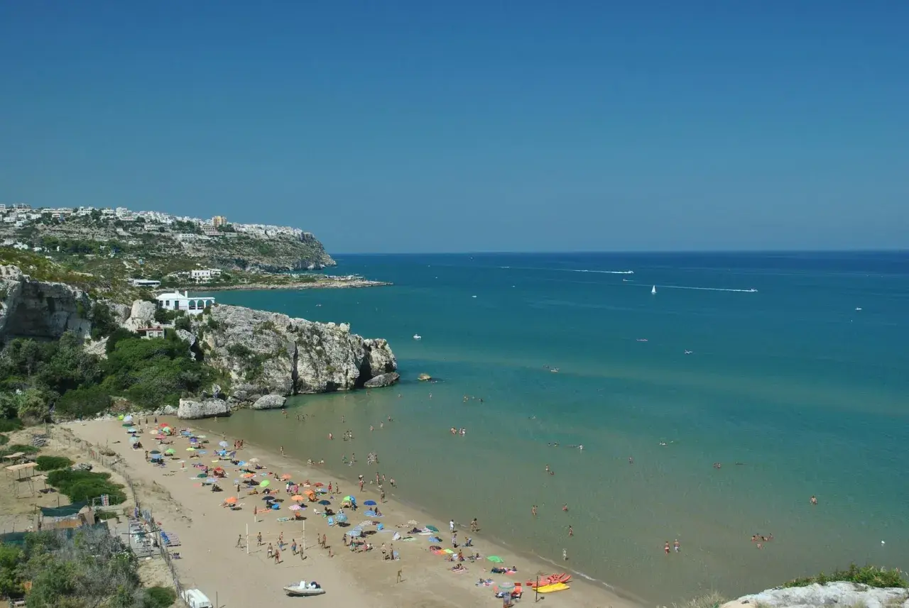 Peschici spiaggia Zaiana, con gente che prende il sole e nuota nelle acque cristalline sotto un cielo azzurro.