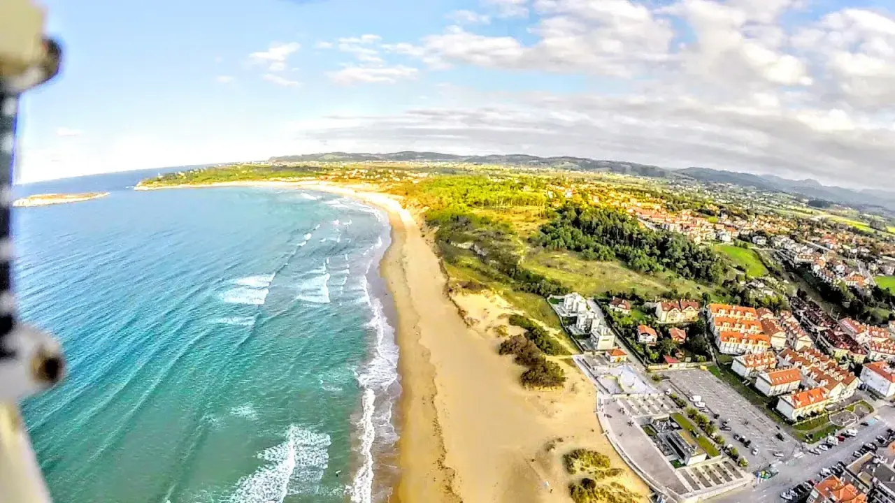 Vista aérea de la playa de Somo, con el mar turquesa, arena dorada, dunas y un pueblo costero con tejados rojos.